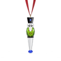 A Swarovski Christmas ornament shaped like a nutcracker, featuring a green body, black top hat, white face, and a silver cone base ending in a blue faceted crystal, suspended from a red ribbon on a white background.